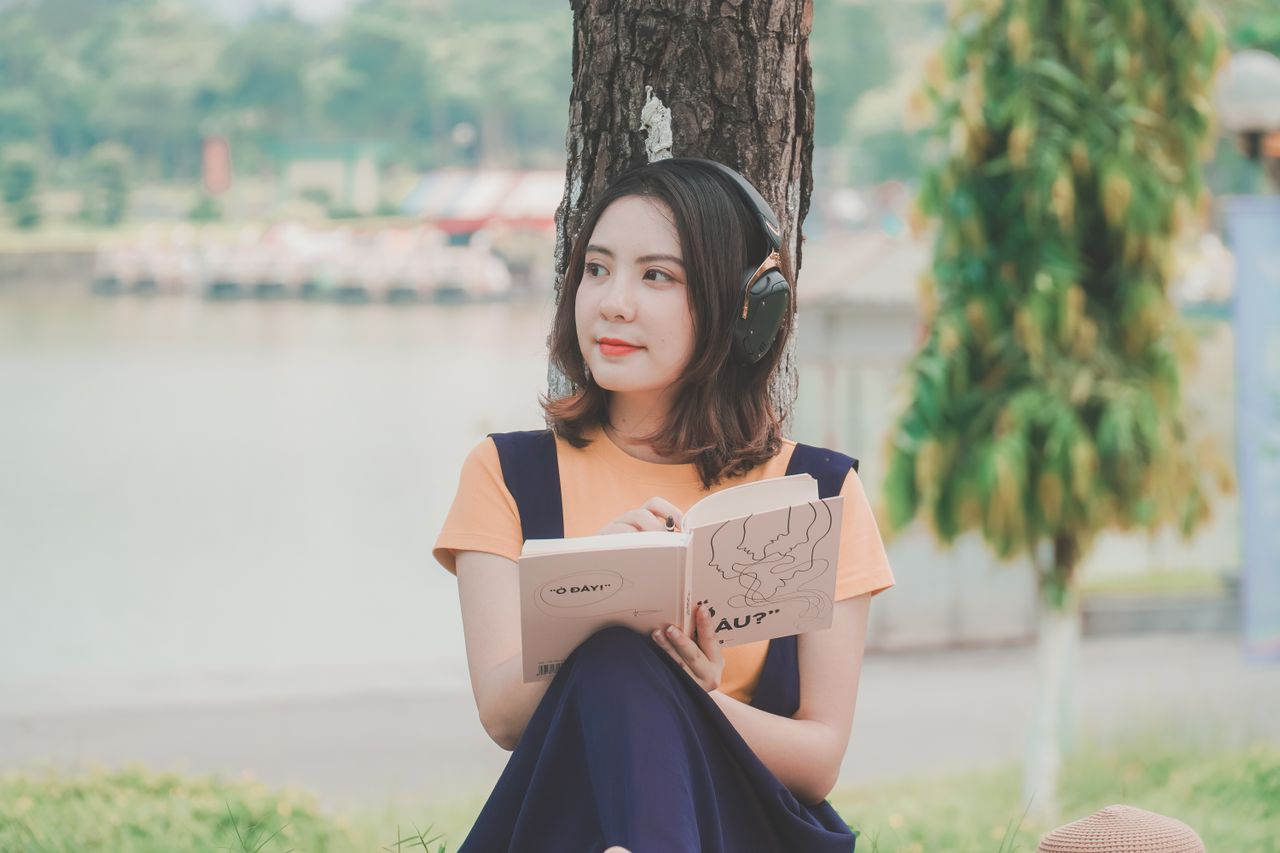 Une jeune femme assise au pied d'un arbre au bord de l'eau, un casque audio sur les oreilles et un carnet ouvert sur les genoux.