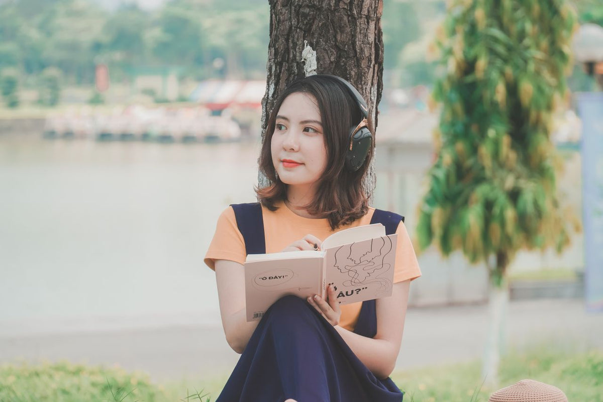 Une jeune femme assise au pied d'un arbre au bord de l'eau, un casque audio sur les oreilles et un carnet ouvert sur les genoux.