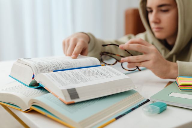 Un adolescent en sweat à capuche, accoudé à une table, feuillette un dictionnaire ouvert posé sur une pile de livres, à côté d'une paire de lunettes, de marque-pages colorés et d'un crayon.
