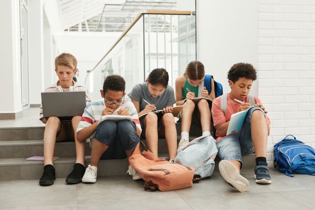 Cinq enfants assis sur des marches, cartables posés à côté d'eux, l'un devant un ordinateur portable, les autres écrivant dans des cahiers.
