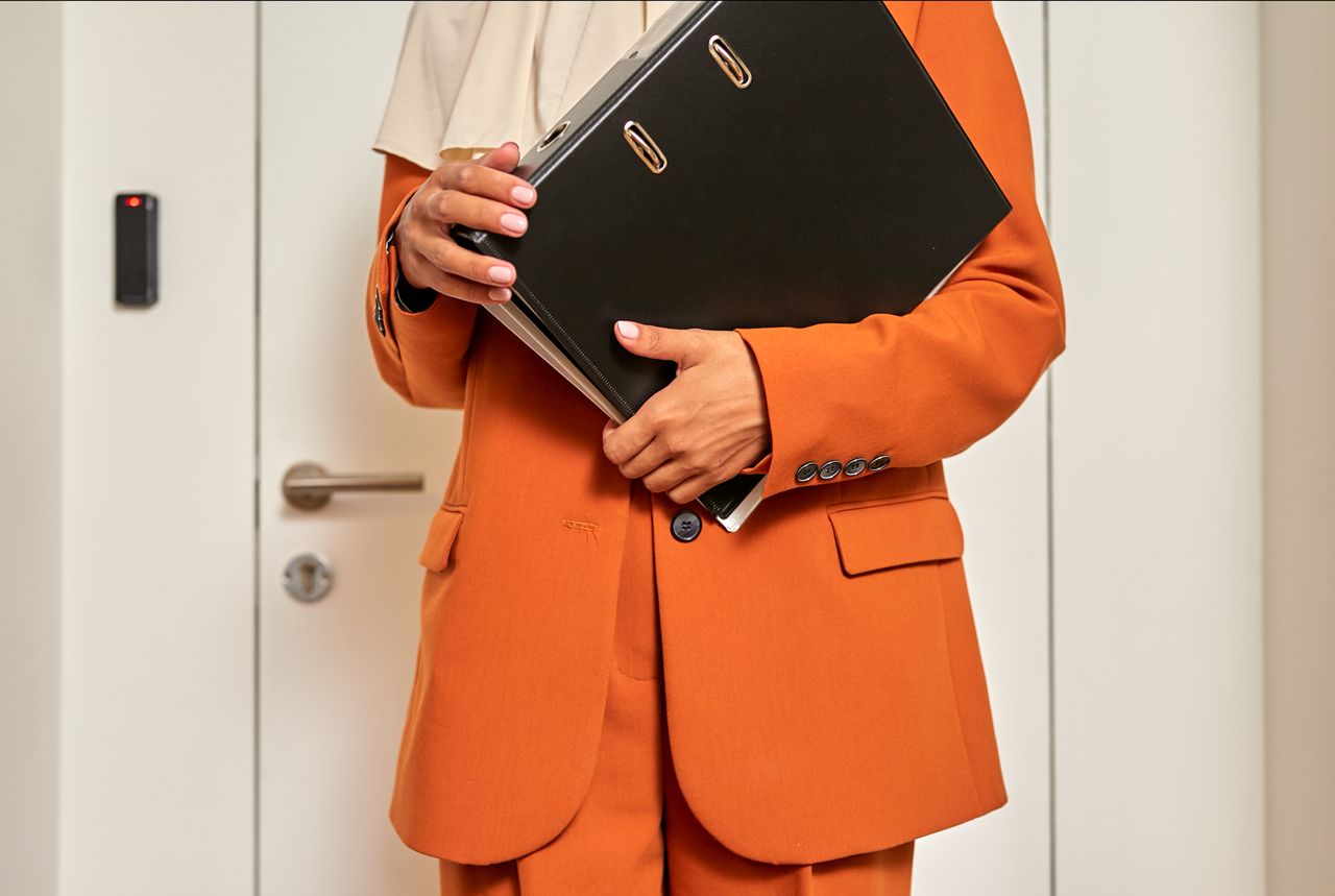 Une personne en veste orange, debout devant une porte, serre un porte-documents noir contre elle.