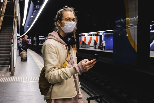 Une personne avec un masque, un casque autour du cou et un sac à dos, consulte son téléphone sur un quai de métro.