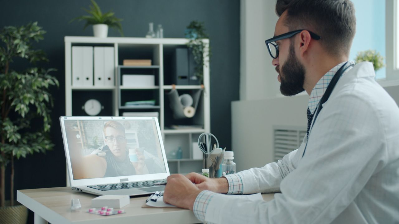 Un soignant en blouse blanche, assis à son bureau, prend des notes pendant une consultation en visioconférence sur son ordinateur portable.