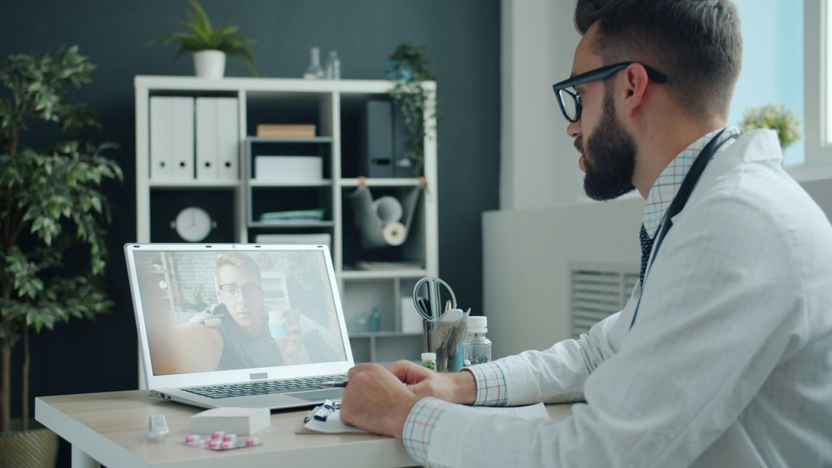 Un soignant en blouse blanche, assis à son bureau, prend des notes pendant une consultation en visioconférence sur son ordinateur portable.