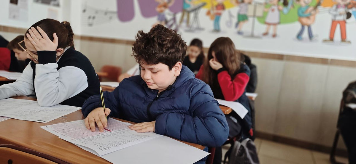 Dans une salle de classe, un élève écrit penché sur son cahier pendant qu'une camarade, derrière lui, se tient la tête à deux mains.
