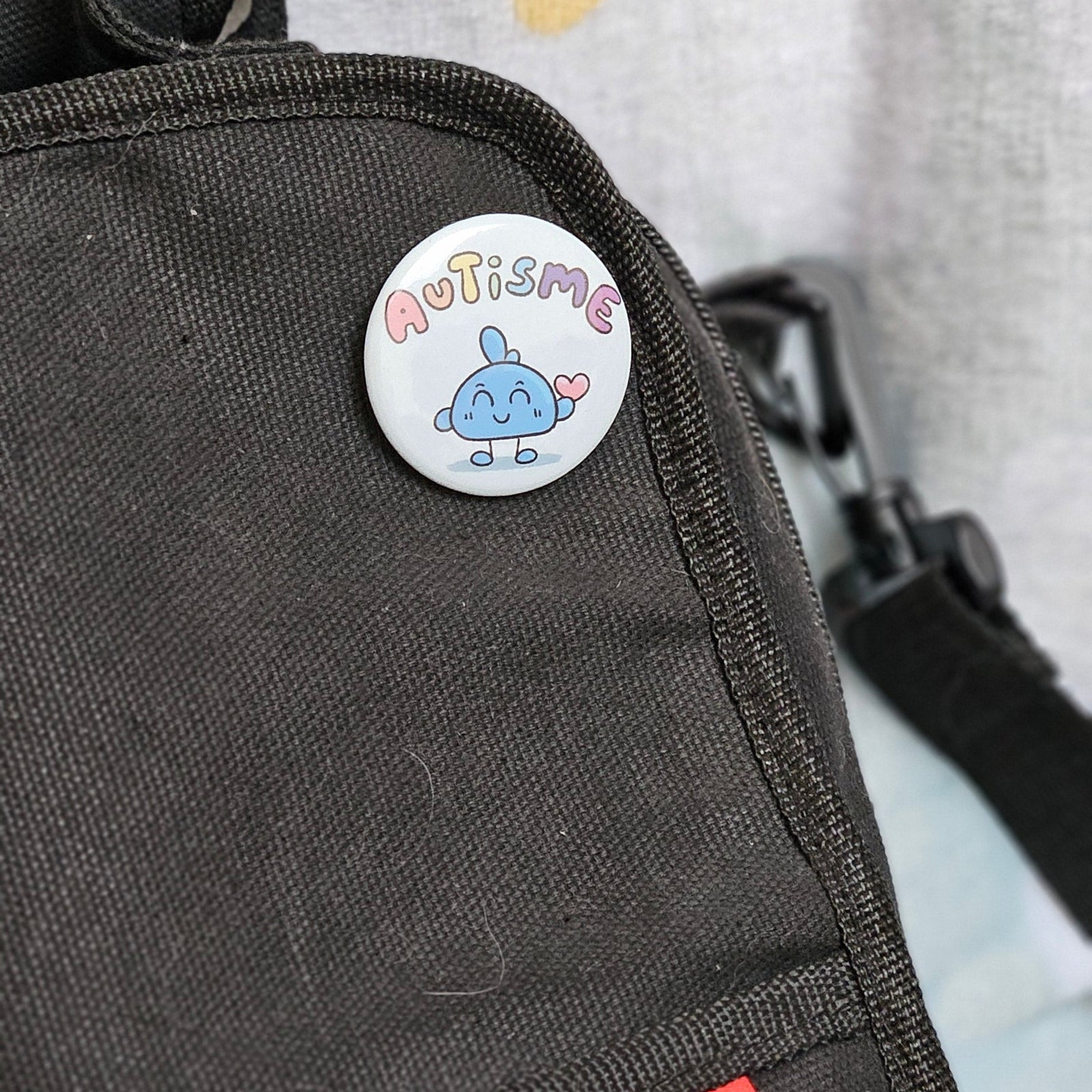 Badge autisme épinglé sur la poche d'un sac en toile gris foncé : petit personnage bleu souriant et un cœur rose sous le mot autisme en lettres pastel