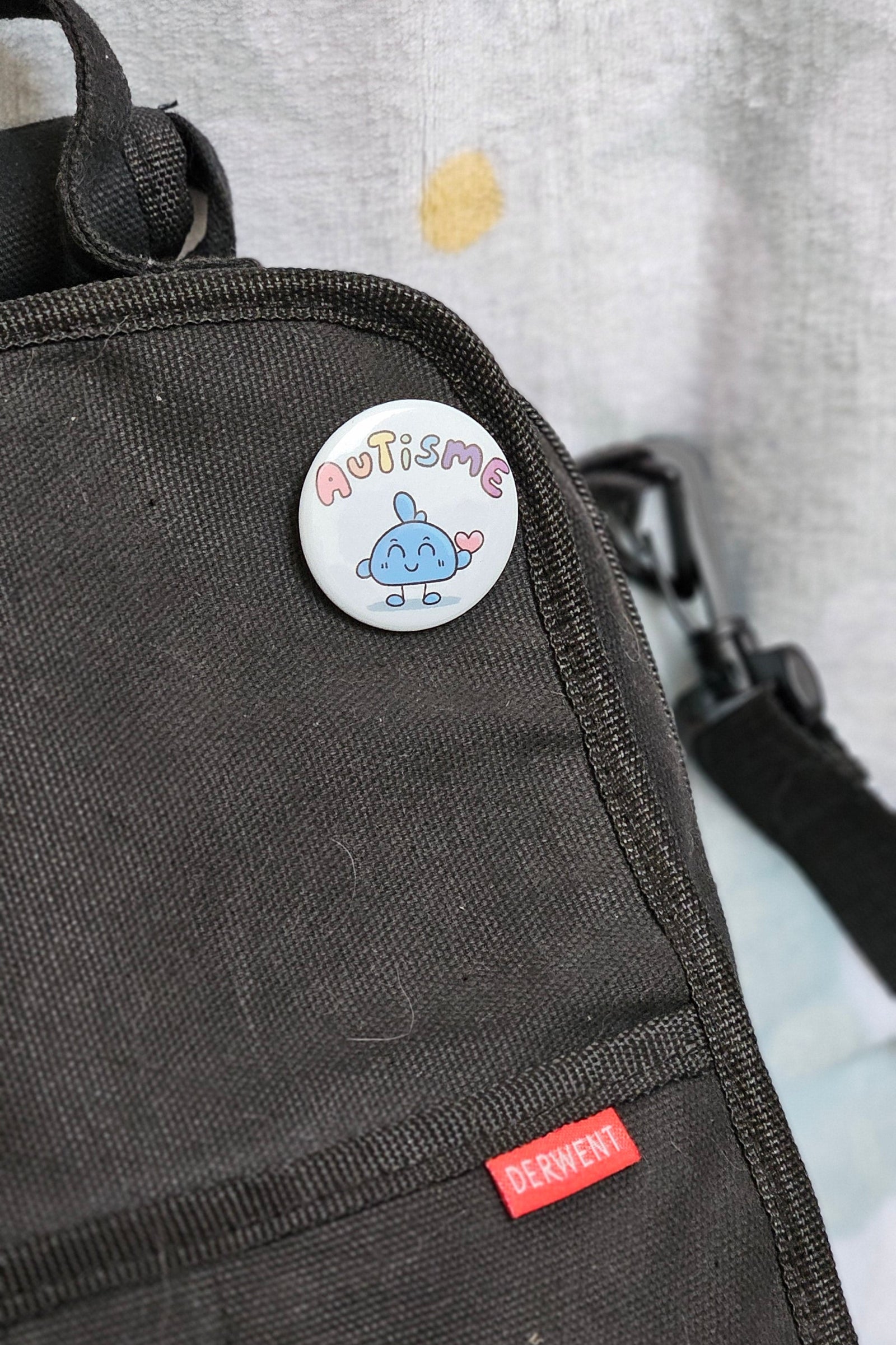 Badge autisme épinglé sur la poche d'un sac en toile gris foncé : petit personnage bleu souriant et un cœur rose sous le mot autisme en lettres pastel