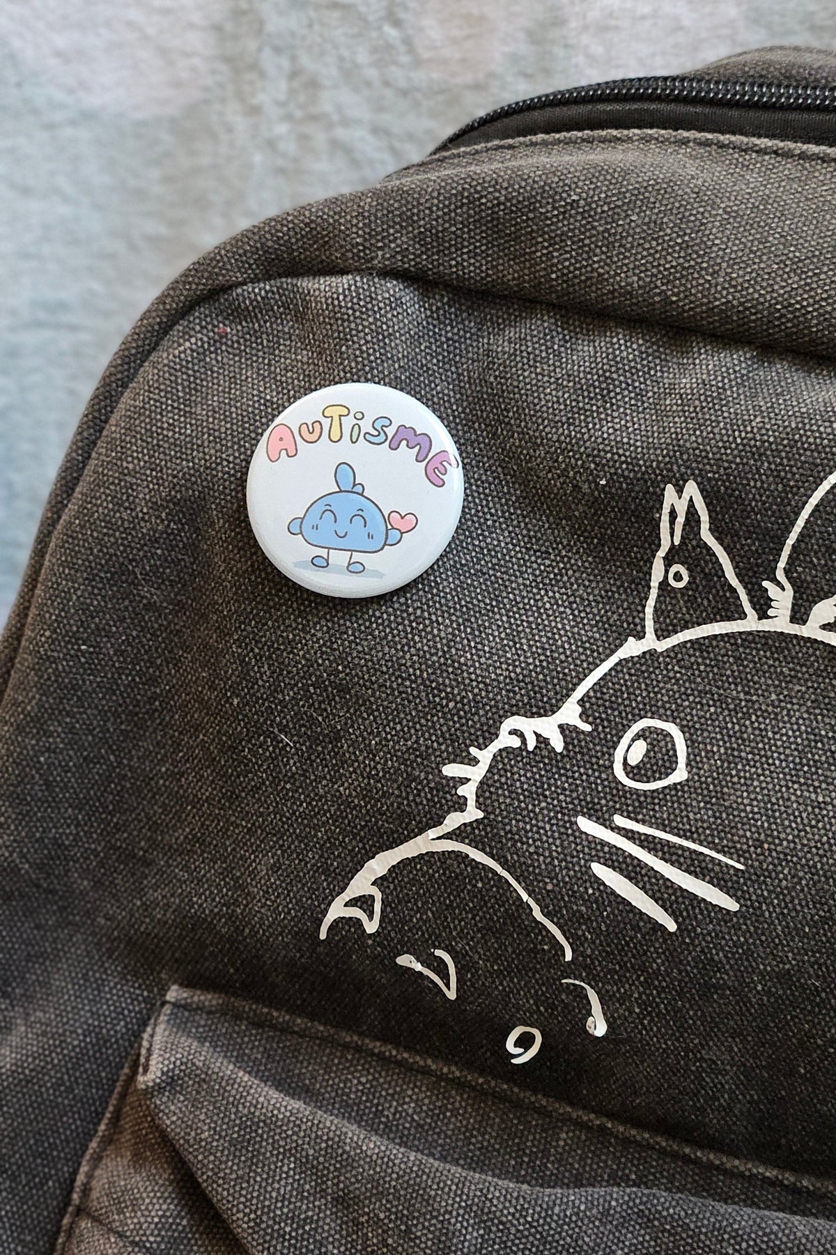 Badge autisme épinglé sur un sac à dos en toile gris foncé décoré d'un dessin au trait blanc