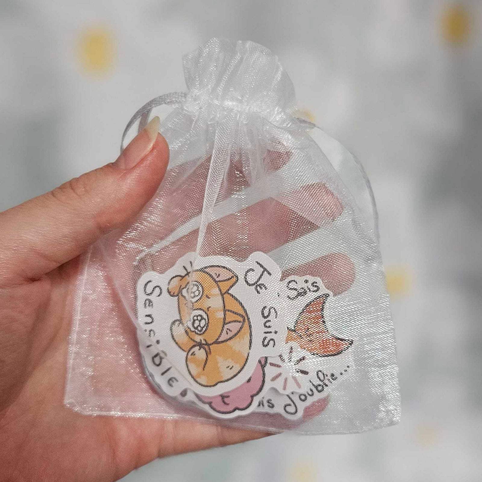 Sachet en organza blanc tenu dans une main, laissant voir des stickers illustrés dont un chat orange endormi portant la mention « Je suis sensible »