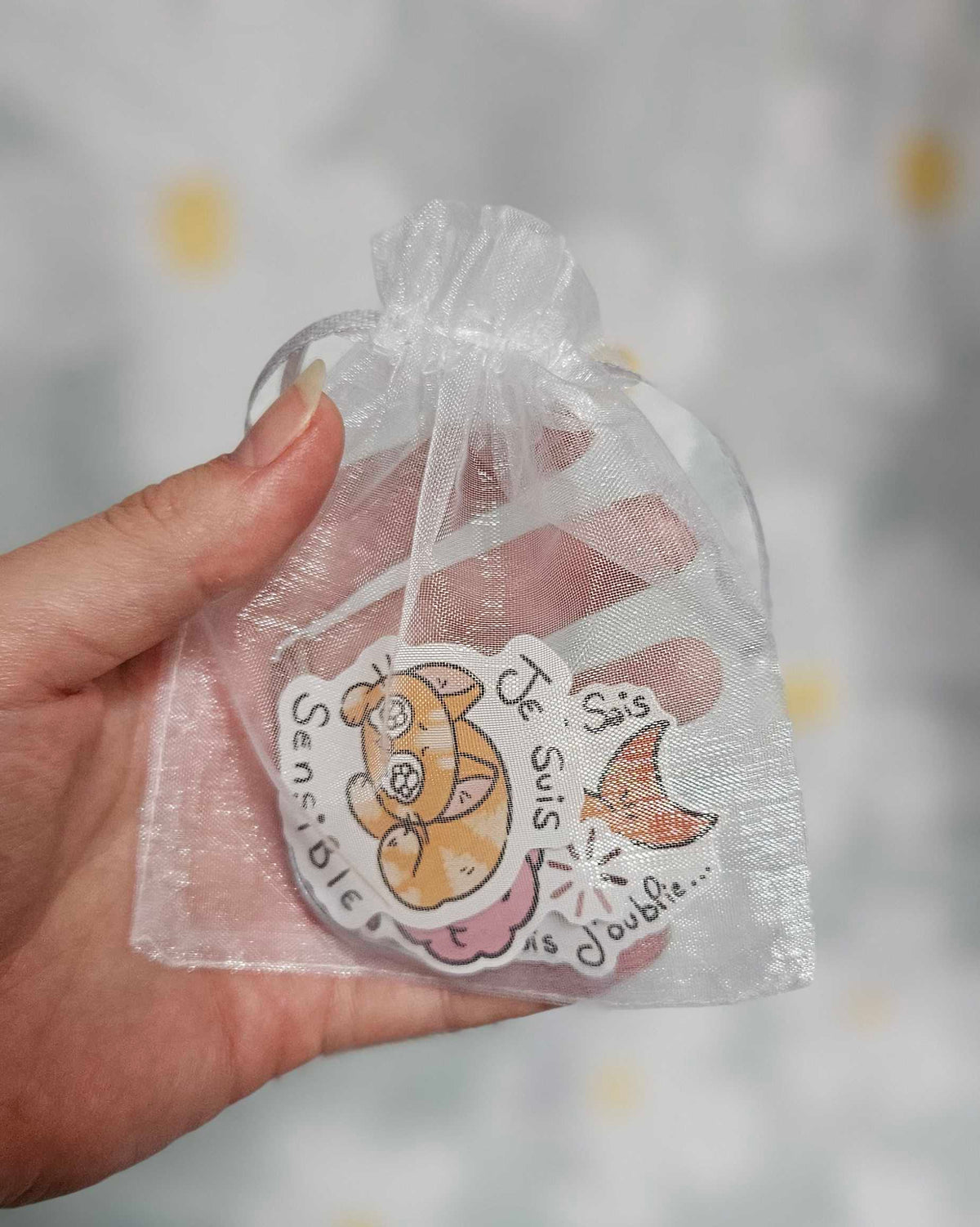 Sachet en organza blanc tenu dans une main, laissant voir des stickers illustrés dont un chat orange endormi portant la mention « Je suis sensible »
