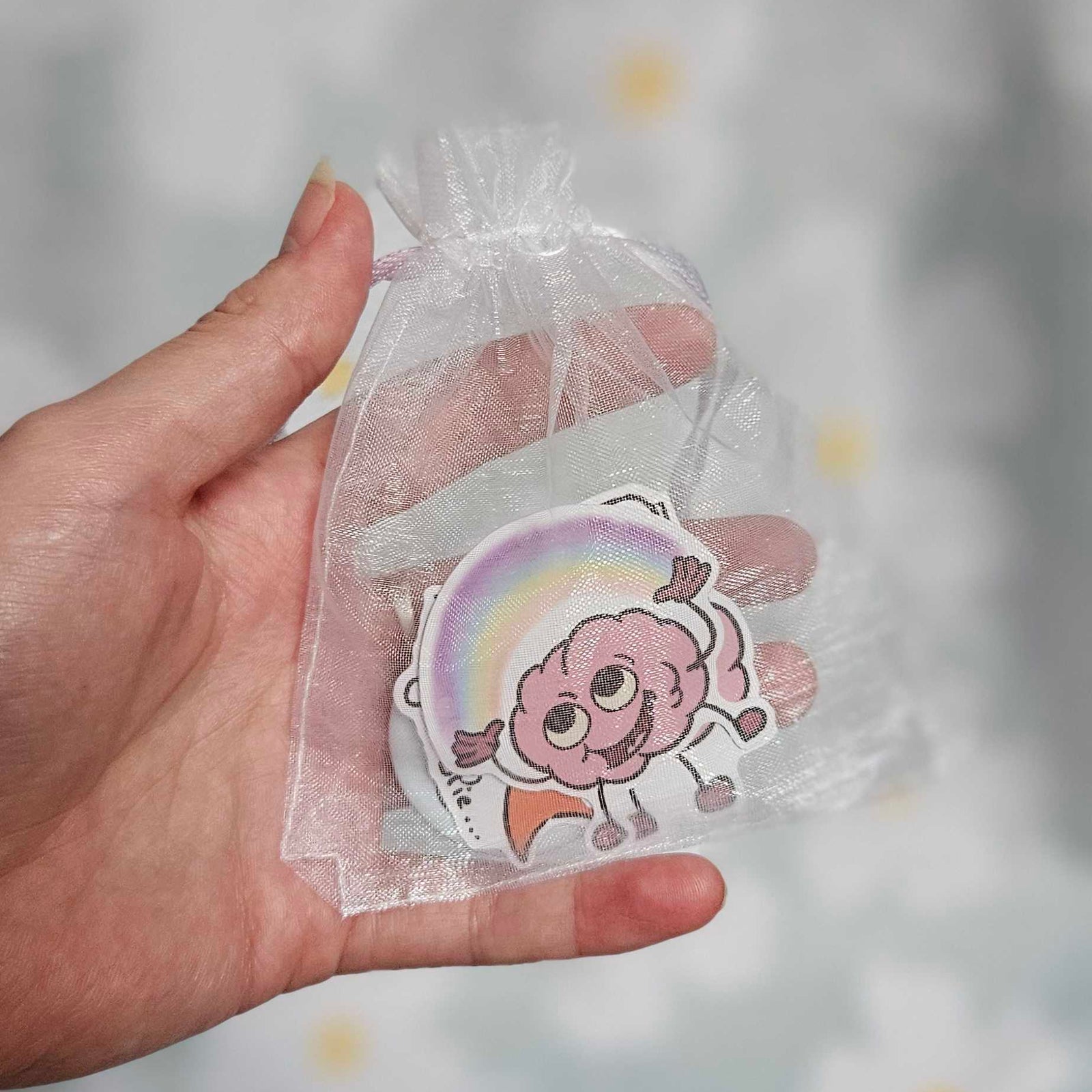 Sachet en organza blanc tenu dans une main, laissant voir un sticker illustré d'un cerveau rose souriant devant un arc-en-ciel