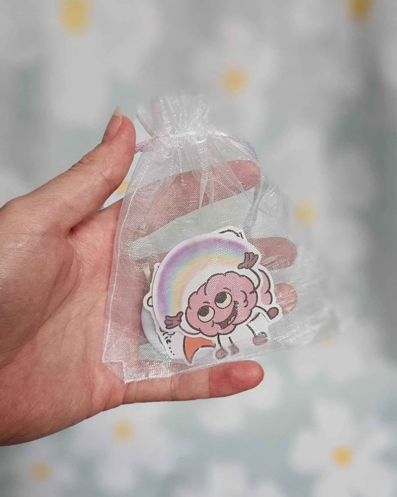 Sachet en organza blanc tenu dans une main, laissant voir un sticker illustré d'un cerveau rose souriant devant un arc-en-ciel