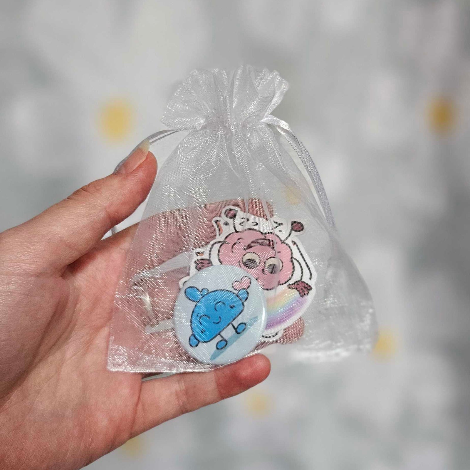 Sachet en organza blanc tenu dans une main, contenant un badge rond bleu et plusieurs stickers illustrés