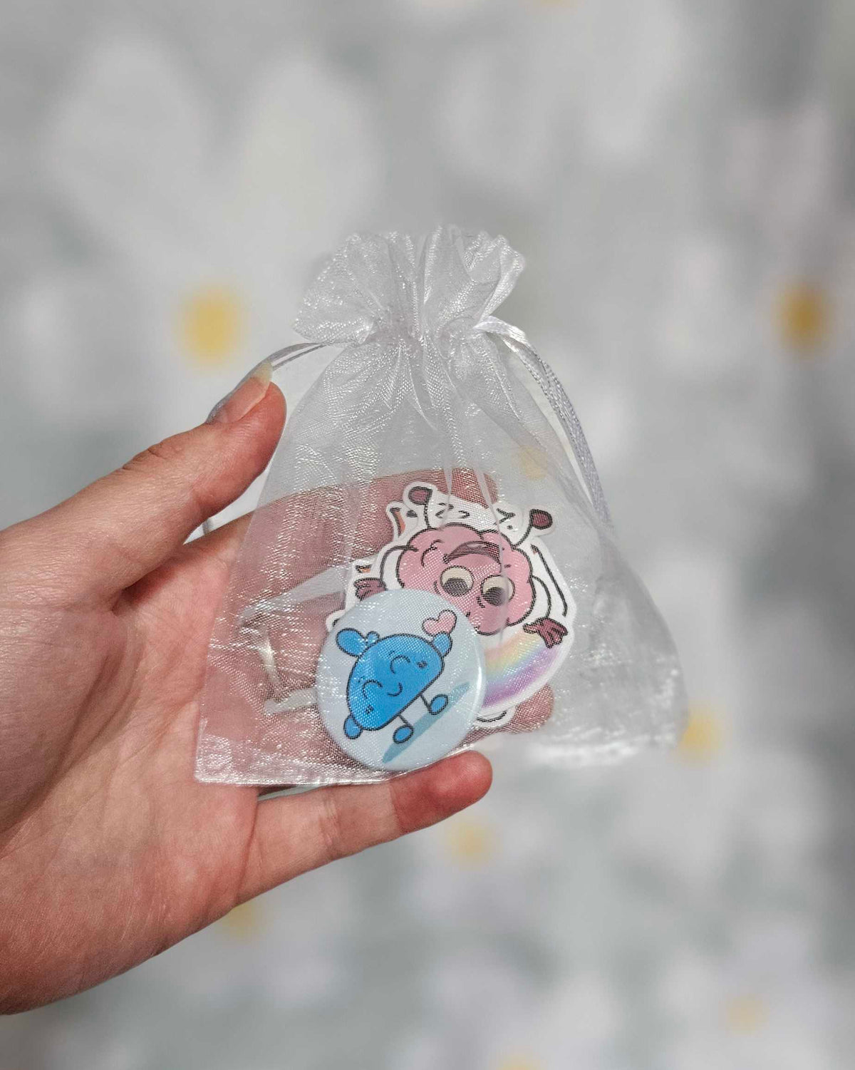 Sachet en organza blanc tenu dans une main, contenant un badge rond bleu et plusieurs stickers illustrés
