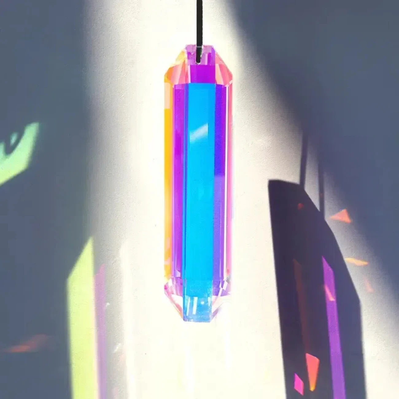 Prisme en cristal suspendu à un fil devant un mur clair traversé par le soleil, ses facettes prenant des teintes bleues, roses et orangées
