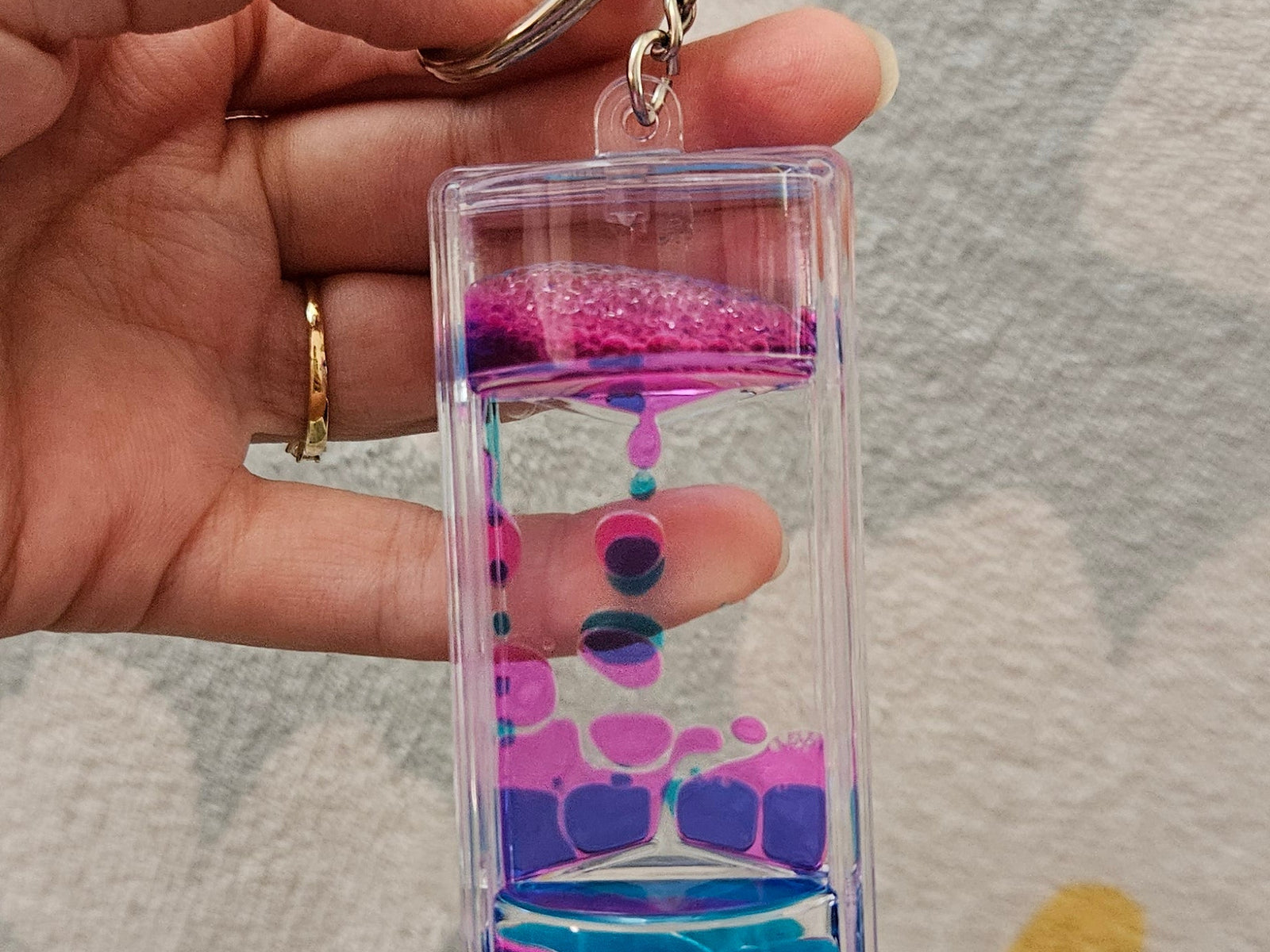Porte-clé minuteur liquide rose et bleu tenu par son anneau, les gouttes colorées descendant lentement dans le boîtier transparent