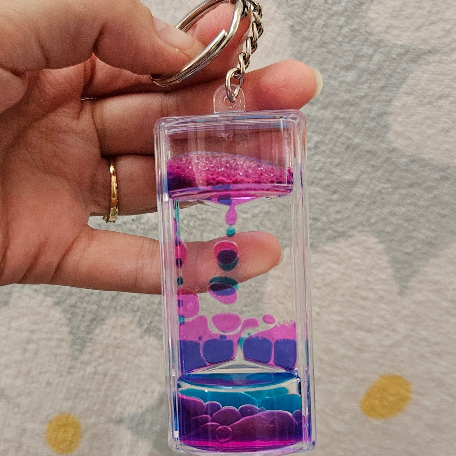 Porte-clé minuteur liquide rose et bleu tenu par son anneau, les gouttes colorées descendant lentement dans le boîtier transparent