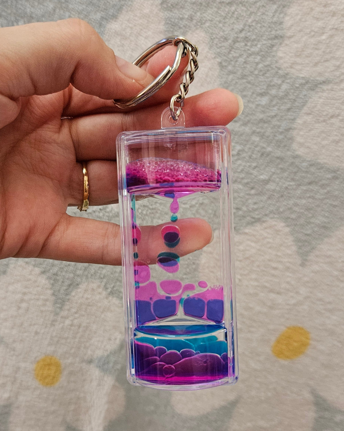 Porte-clé minuteur liquide rose et bleu tenu par son anneau, les gouttes colorées descendant lentement dans le boîtier transparent