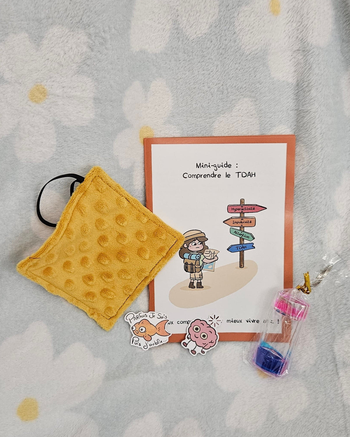 Contenu du Kit TDAH posé à plat sur un plaid fleuri : le mini-guide Comprendre le TDAH, un Mini SAM Bubble moutarde, un porte-clé minuteur liquide rose et bleu, et deux stickers, le poisson « Parfois je sais, puis j'oublie » et le cerveau en chargement