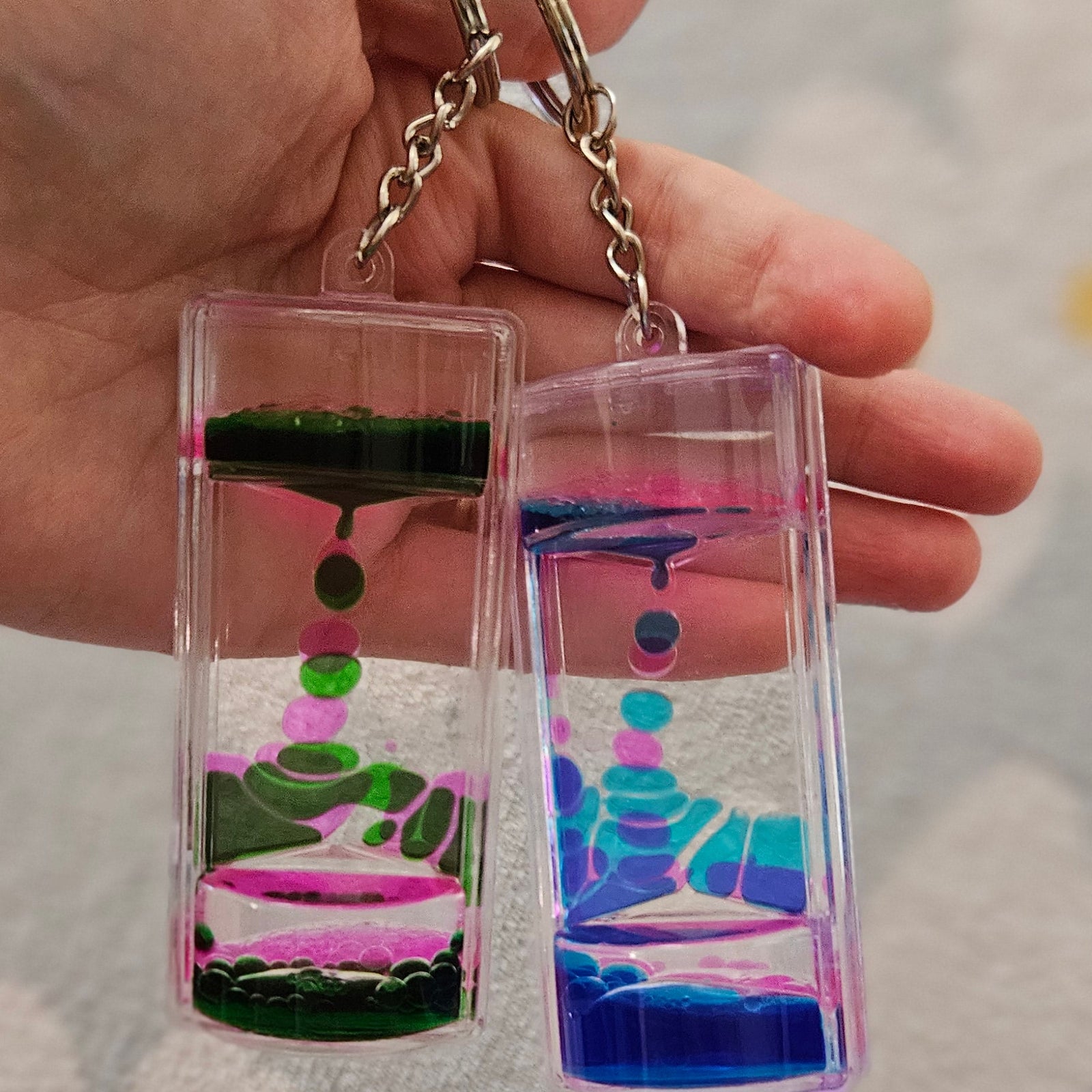 Deux porte-clés minuteurs liquides tenus côte à côte dans une main, l'un aux gouttes vertes et roses, l'autre aux gouttes bleues et turquoise