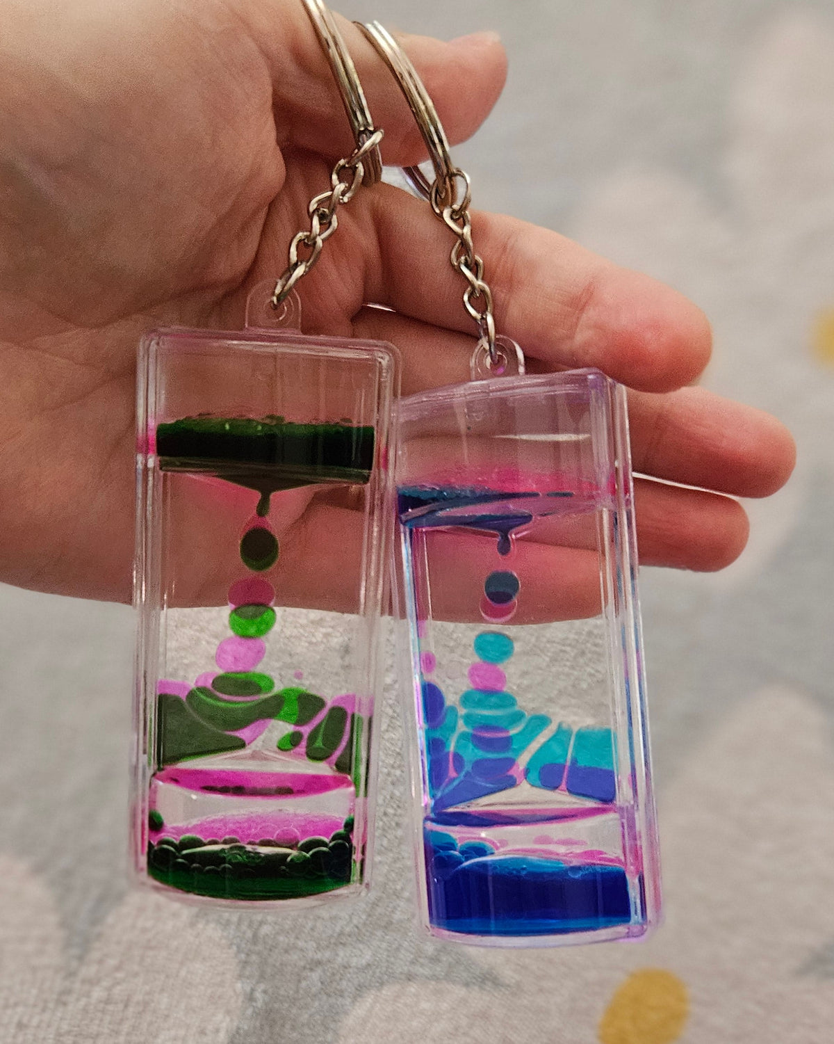 Deux porte-clés minuteurs liquides tenus côte à côte dans une main, l'un aux gouttes vertes et roses, l'autre aux gouttes bleues et turquoise