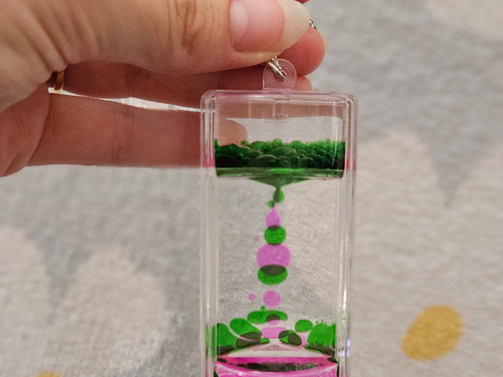 Porte-clé minuteur liquide vert et rose tenu par son anneau, les gouttes colorées descendant lentement dans le boîtier transparent