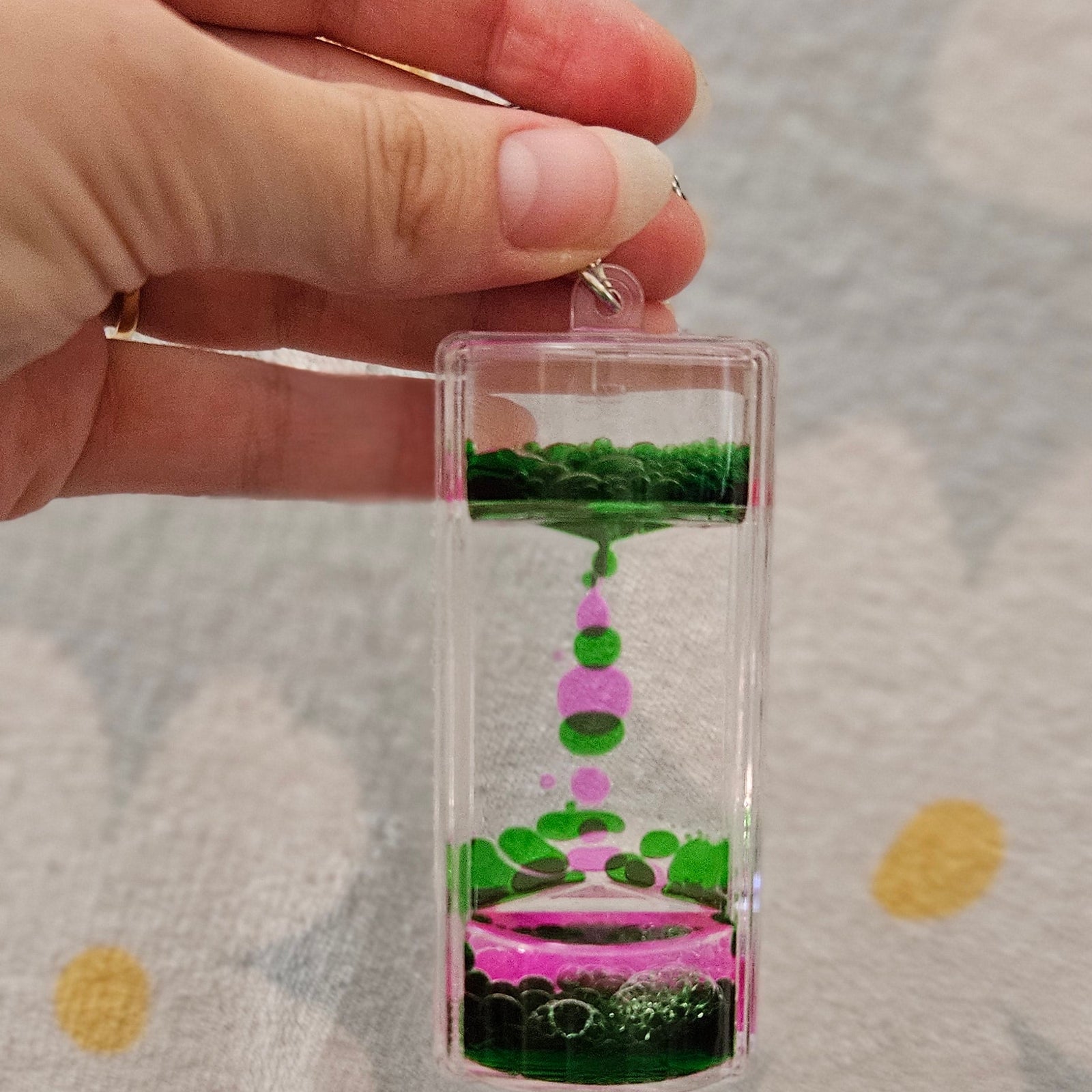 Porte-clé minuteur liquide vert et rose tenu par son anneau, les gouttes colorées descendant lentement dans le boîtier transparent