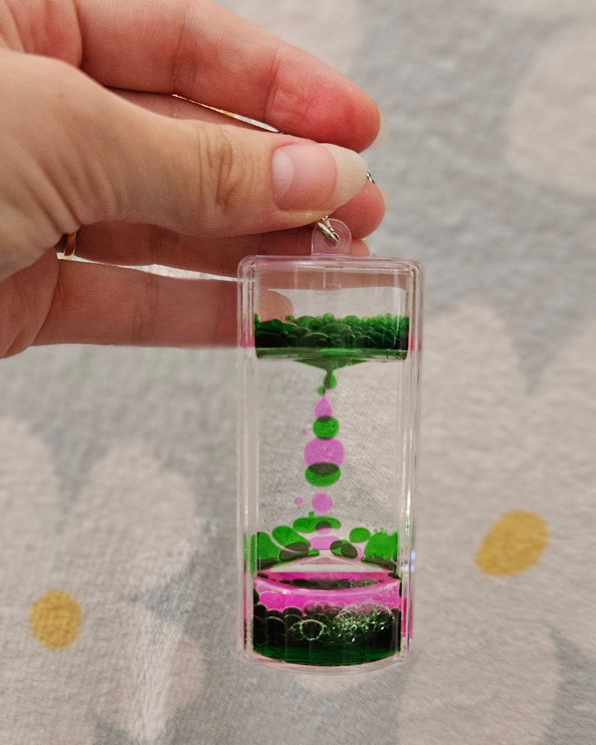 Porte-clé minuteur liquide vert et rose tenu par son anneau, les gouttes colorées descendant lentement dans le boîtier transparent